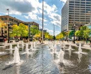 A fountain in the middle of a plaza with trees and buildings in the background.