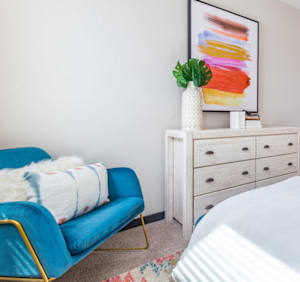 A blue chair with a white cushion is in front of a white dresser with a plant on top.