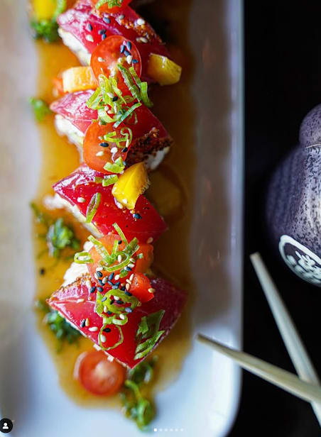 A dish of sliced red fish topped with green herbs and seeds.
