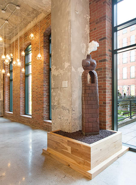 A large planter with a brick chimney on top is in the middle of a room with brick walls and windows.