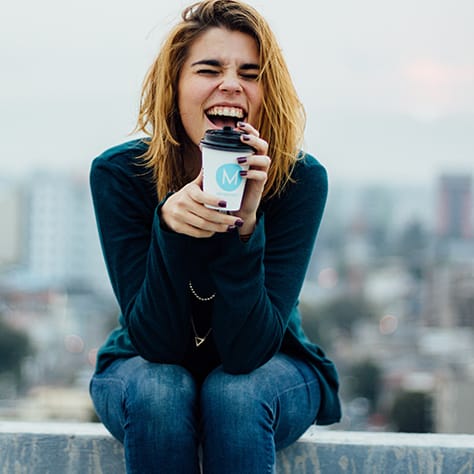 A women sitting on a rooftop ledge drinking coffee.