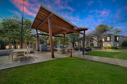 A wooden pavilion is surrounded by a grassy area and a patio with chairs.