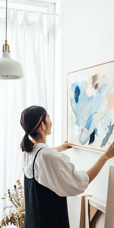 A woman is hanging a painting on a white wall.