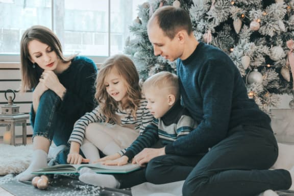 a family sitting on the floor in front of a christmas tree reading a book