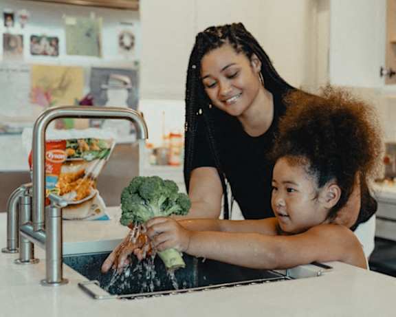 a woman and a child washing vegetables in the kitchen sink