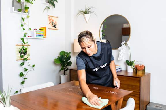 A woman in a black Spruce t-shirt is cleaning a wooden table with a green cloth.