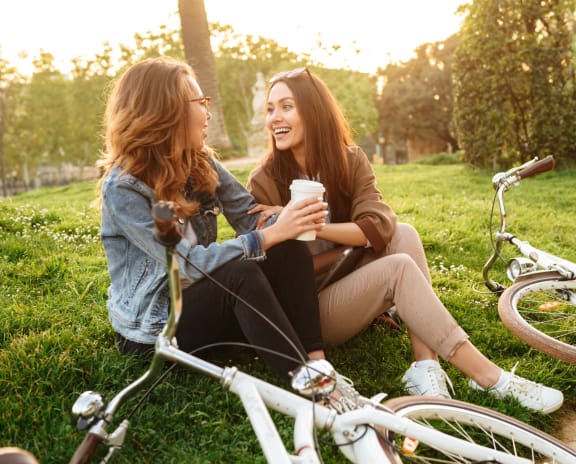 Two Women Sitting In Grass Having Conversation Next to Their Bikes 