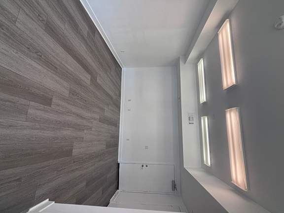 Live Work Apartments for Rent in Corona, CA - Dos Lagos Townhomes - Empty Room With Gray Wood-Style Flooring, White Walls, Overhead Fluorescent Lighting, And A Closed Door.