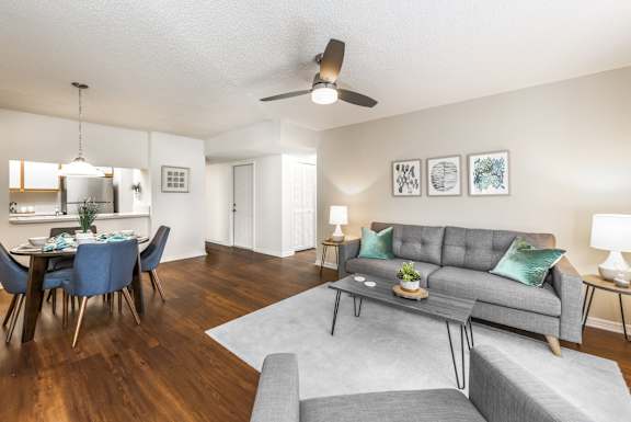 Livingroom with wood like style floors, couch, ceiling fan, arm chair, end tables, table lamps, area rug, dining area with four chairs and handing light and the kitchen in the background.