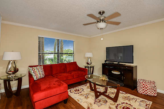 Sectional couch, area rug, TV stand, large TV, end table, table lamp, ceiling fan and wood like style floors.