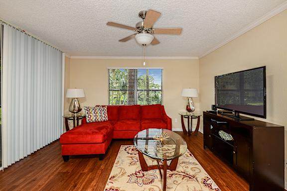 Living room with coffee table, area rug, sectional couch, two end tables, two table lamps, tv stand, large TV, fan and wood like style flooring.
