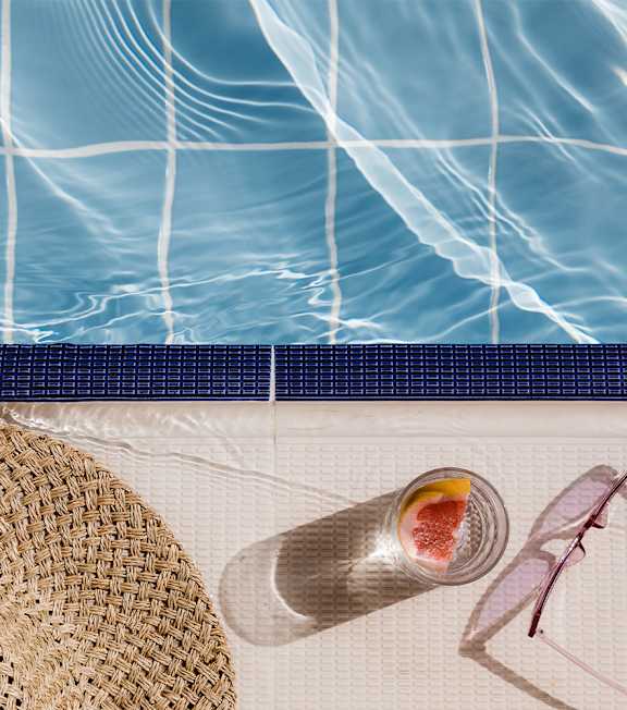 A glass of water with a slice of grapefruit sits on the edge of a pool.