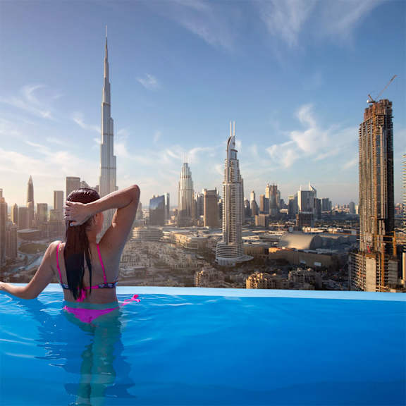 A woman in a pink bikini relaxes in a swimming pool overlooking a city skyline.