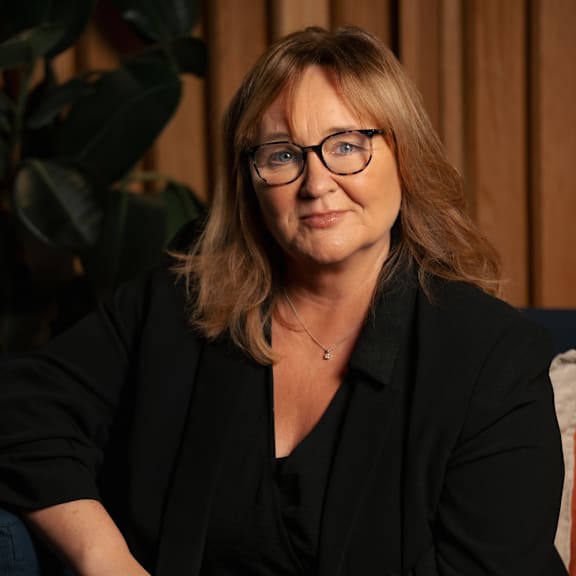 A woman with glasses and a black shirt is sitting in front of a plant.
