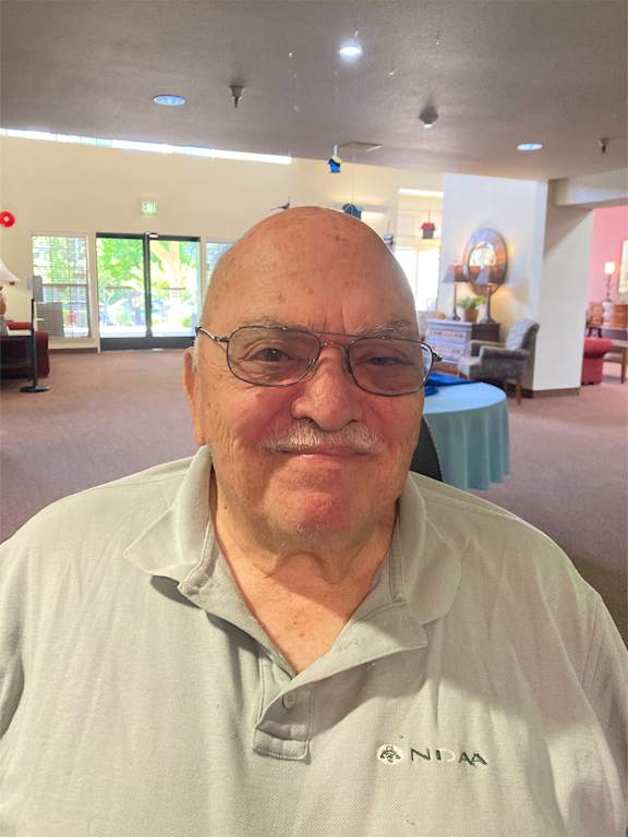 An elderly man with glasses and a striped shirt is smiling at the camera.