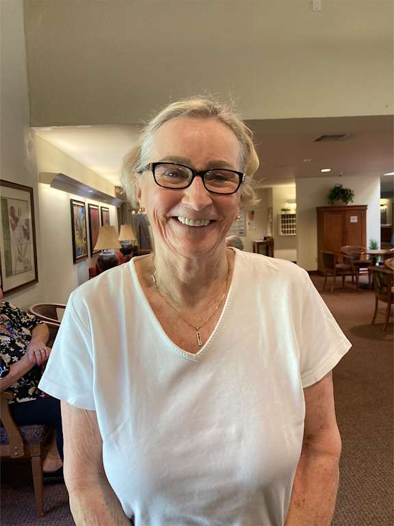 A woman with glasses and a white shirt is smiling in a room with furniture and artwork.