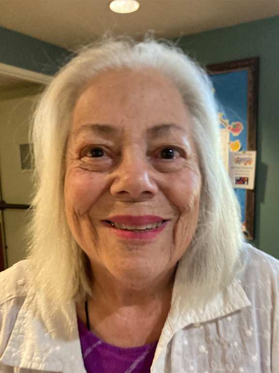 A woman with white hair and a purple shirt is smiling.