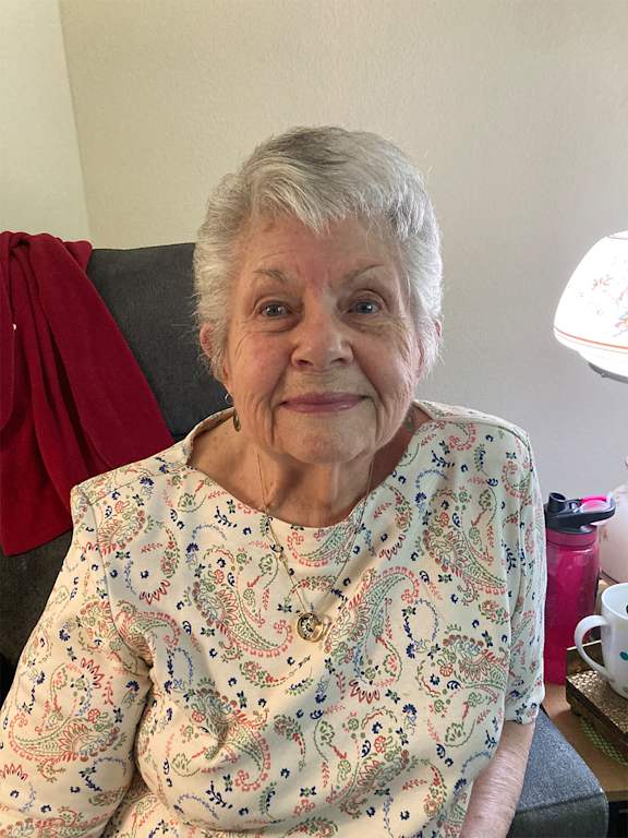An elderly woman with a floral shirt sits in a chair.