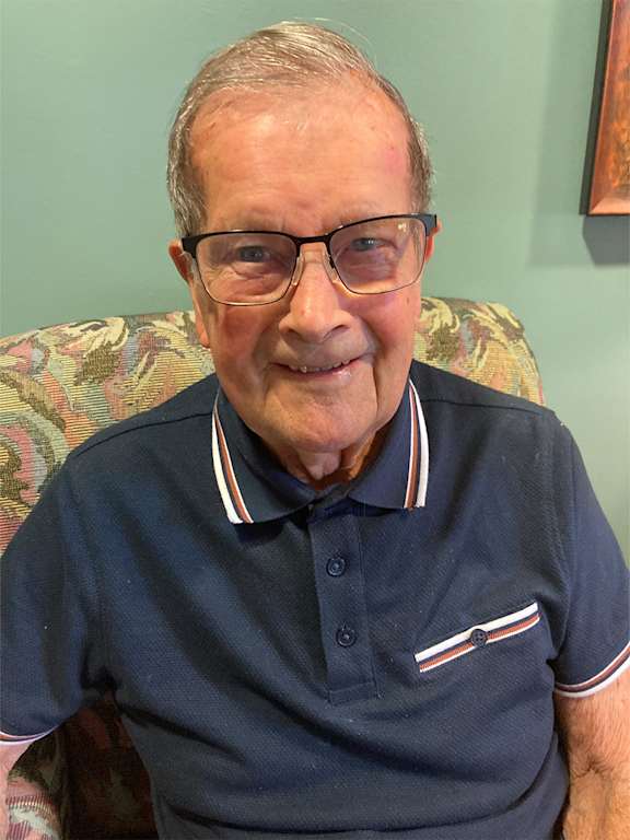 An elderly man with glasses and a striped shirt is smiling at the camera.
