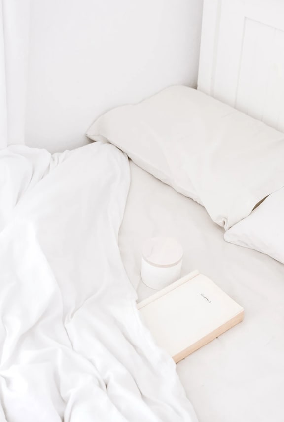 an unmade bed with white sheets and a book and a coffee cup on it