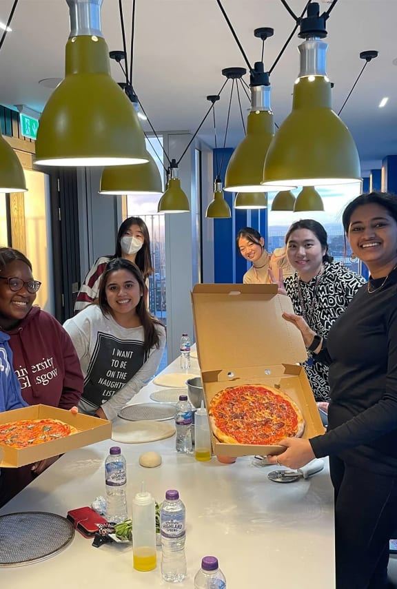 a group of people standing around a table with a box of pizza