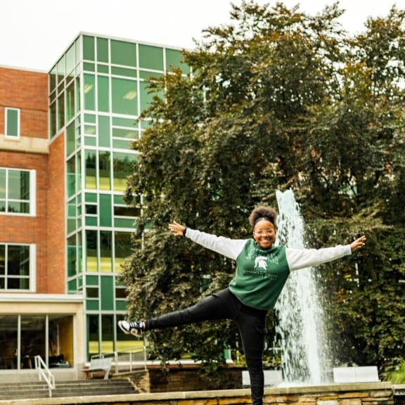 a woman jumping on a ledge in front of a fountain