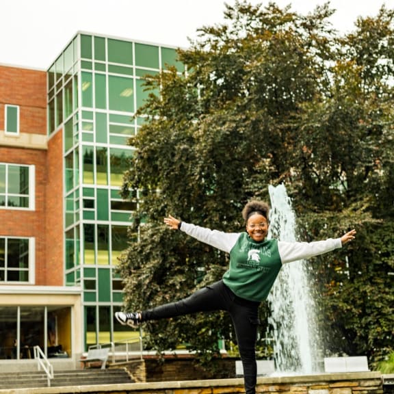 a woman jumping on a ledge in front of a fountain