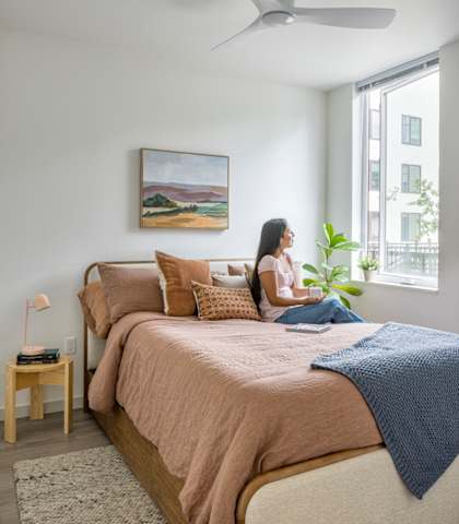 A woman is sitting on a bed with a blanket, a lamp, and a painting on the wall.