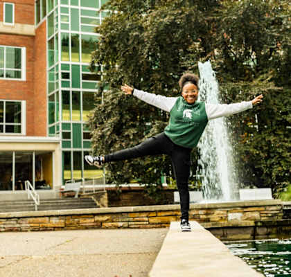 a woman jumping on a ledge in front of a fountain