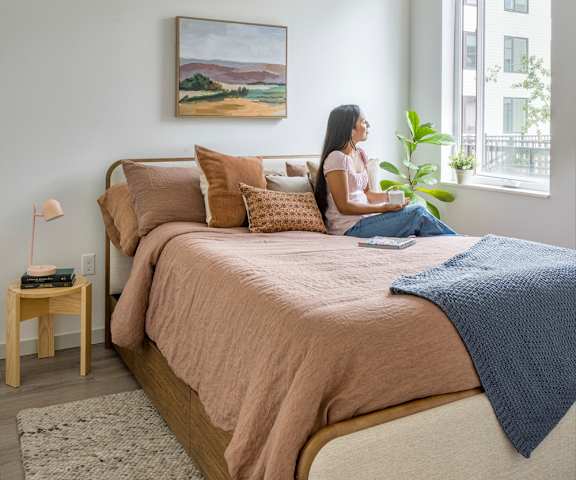 A woman is sitting on a bed with a blanket, a lamp, and a painting on the wall.