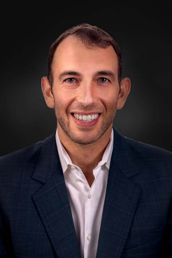 A man in a blue checkered blazer and white shirt smiles at the camera.