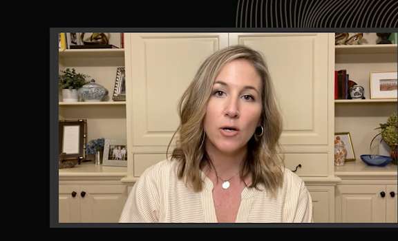 A woman with blonde hair and a white cardigan is speaking in front of a shelf with various decorative items.