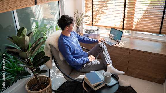 A man in a blue shirt is sitting on a chair with a laptop on his lap.
