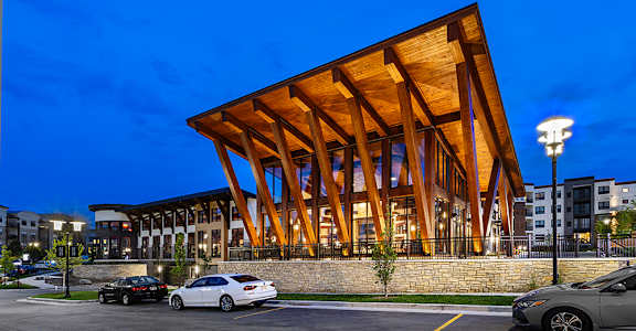 A modern building with a wooden facade is illuminated at dusk with cars parked in front.