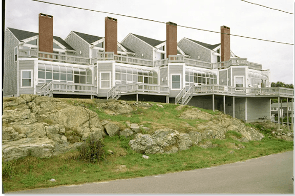 A large white house with a grey roof and a stone wall in front.