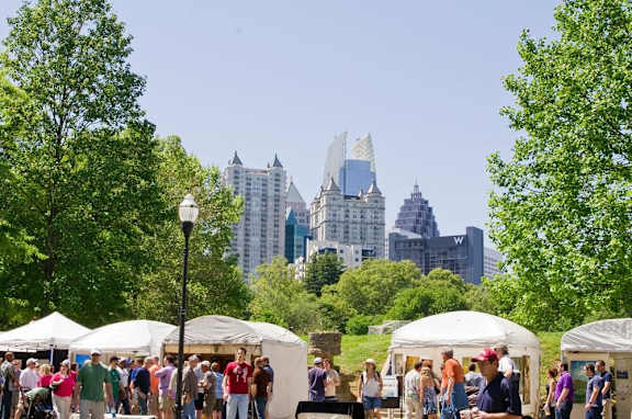A group of people are gathered in a park with white tents and trees.