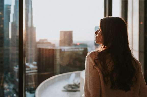 A woman in a beige sweater is looking out a window at a cityscape.