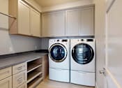 Thumbnail 5 of 13 - A laundry room with a front loading washer and dryer, overhead storage cabinets, and a built in countertop with a sink.