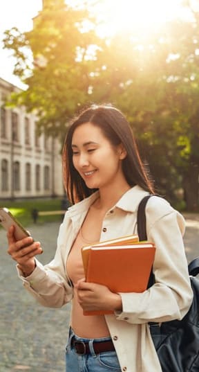 a woman walking down the street looking at her cell phone and holding a book