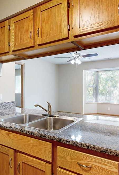 Open kitchen counter with double sink, wood cabinets, and breakfast bar connecting to a bright living area with bay window.