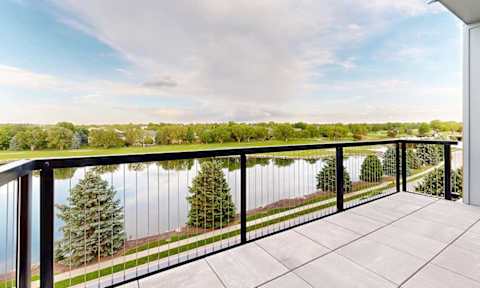 A large balcony in a penthouse apartment overlooks a body of water with trees in the distance.