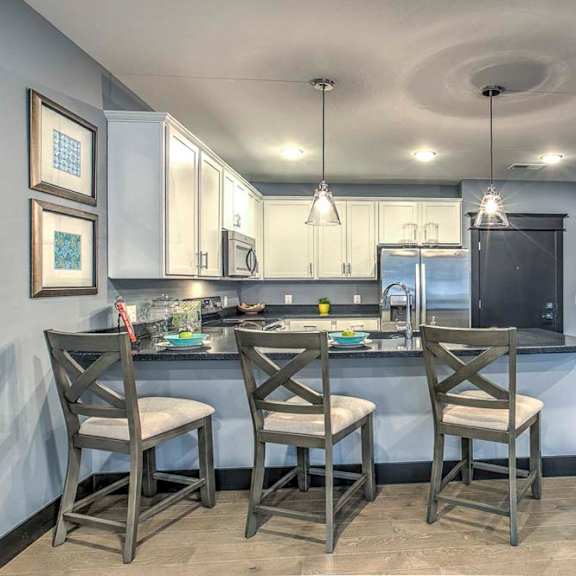 Modern apartment kitchen with white cabinetry, stainless steel appliances, and a dark countertop island with three bar stools and pendant lighting above.