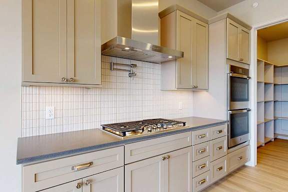 A close-up of the gourmet kitchen with a gas cooktop, sleek vent hood, double wall ovens, tile backsplash, and soft taupe cabinetry with brushed nickel hardware.