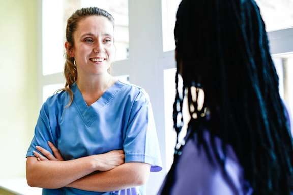 A woman in a blue scrub is standing with her arms crossed while talking to a woman with long black hair.