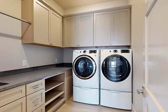 A laundry room with a front loading washer and dryer, overhead storage cabinets, and a built in countertop with a sink.