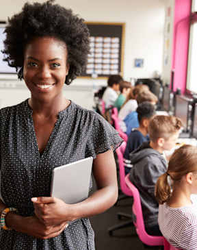 A woman is standing in front of a computer lab with students working on computers.