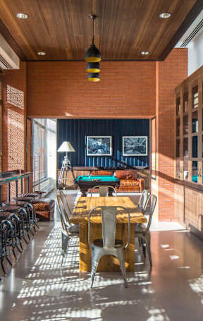 A long wooden table is surrounded by chairs in a room with a brick wall and a long window at The Maximilian Apartments, Long Island City, NY