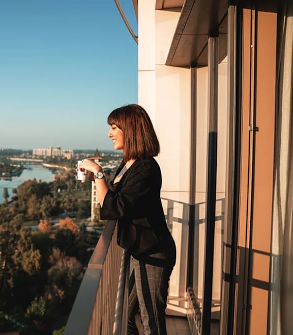 A woman is standing on a balcony with a camera, looking out at a cityscape.