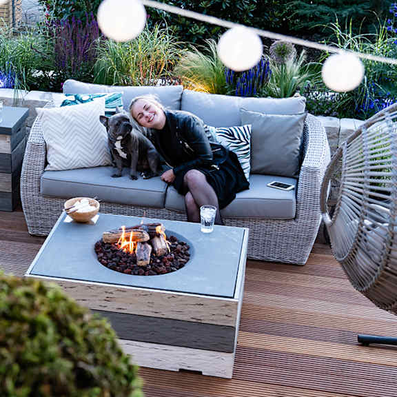 A woman is sitting on a grey couch with a dog beside her, in front of a fire pit.