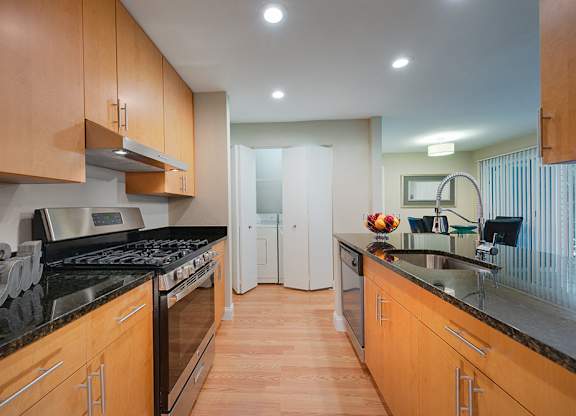 Kitchen with black granite countertops, stainless steel gas stove, upper and lower cabinets, and plank flooring. Double doors in background show washer and dryer.